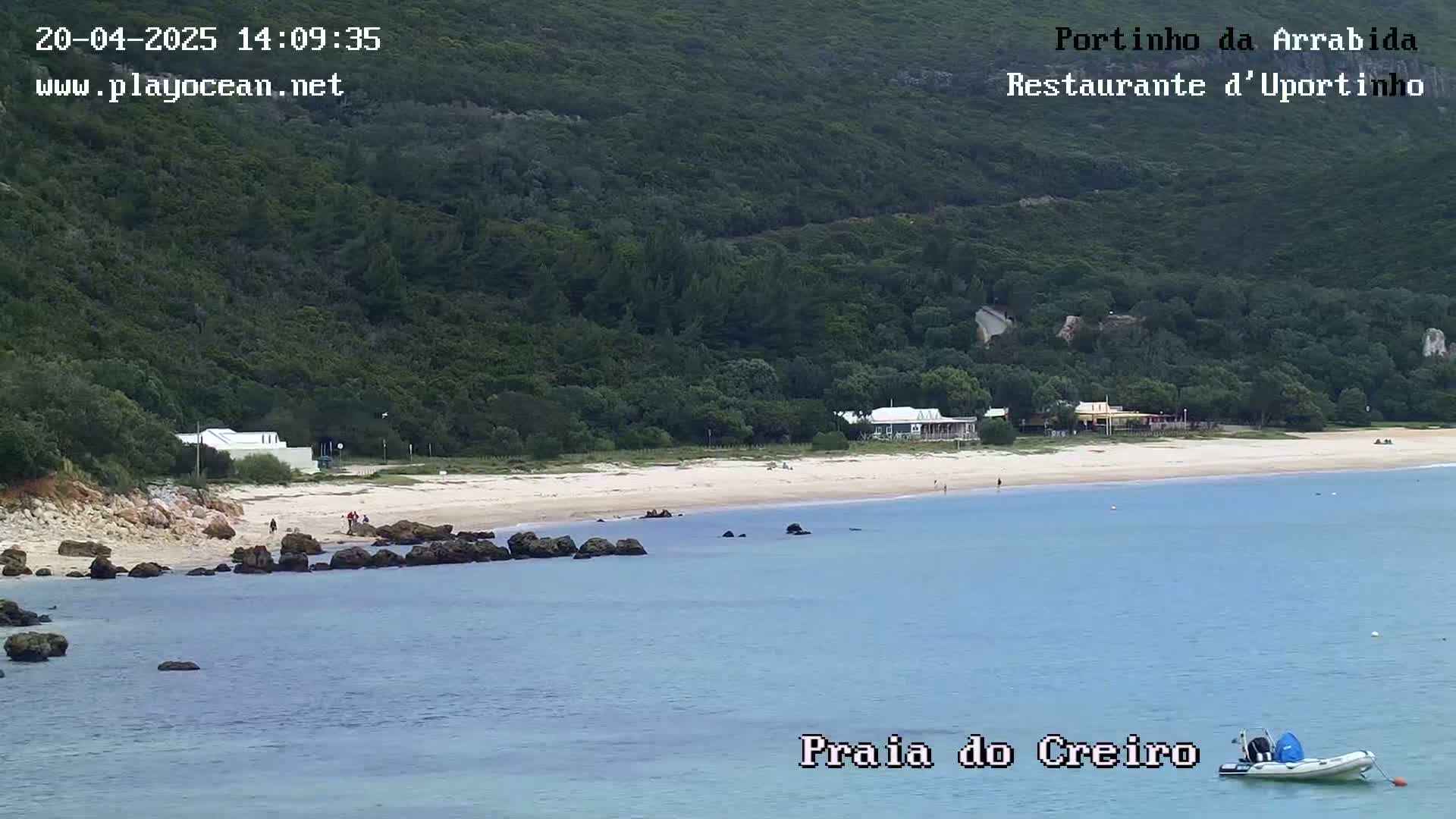 A sandy beach with a few people and buildings is nestled below a lush green hillside, overlooking calm blue water and a small inflatable boat.