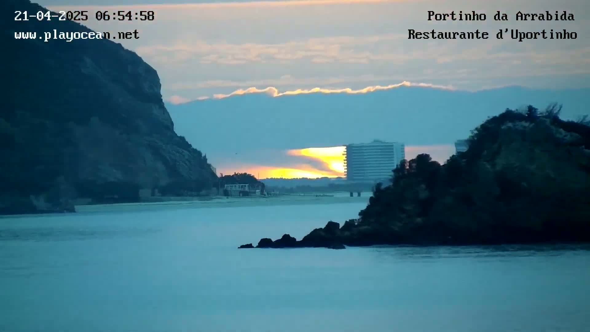 A sunrise illuminates a calm bay between two rocky hills, with a building visible in the distance.
