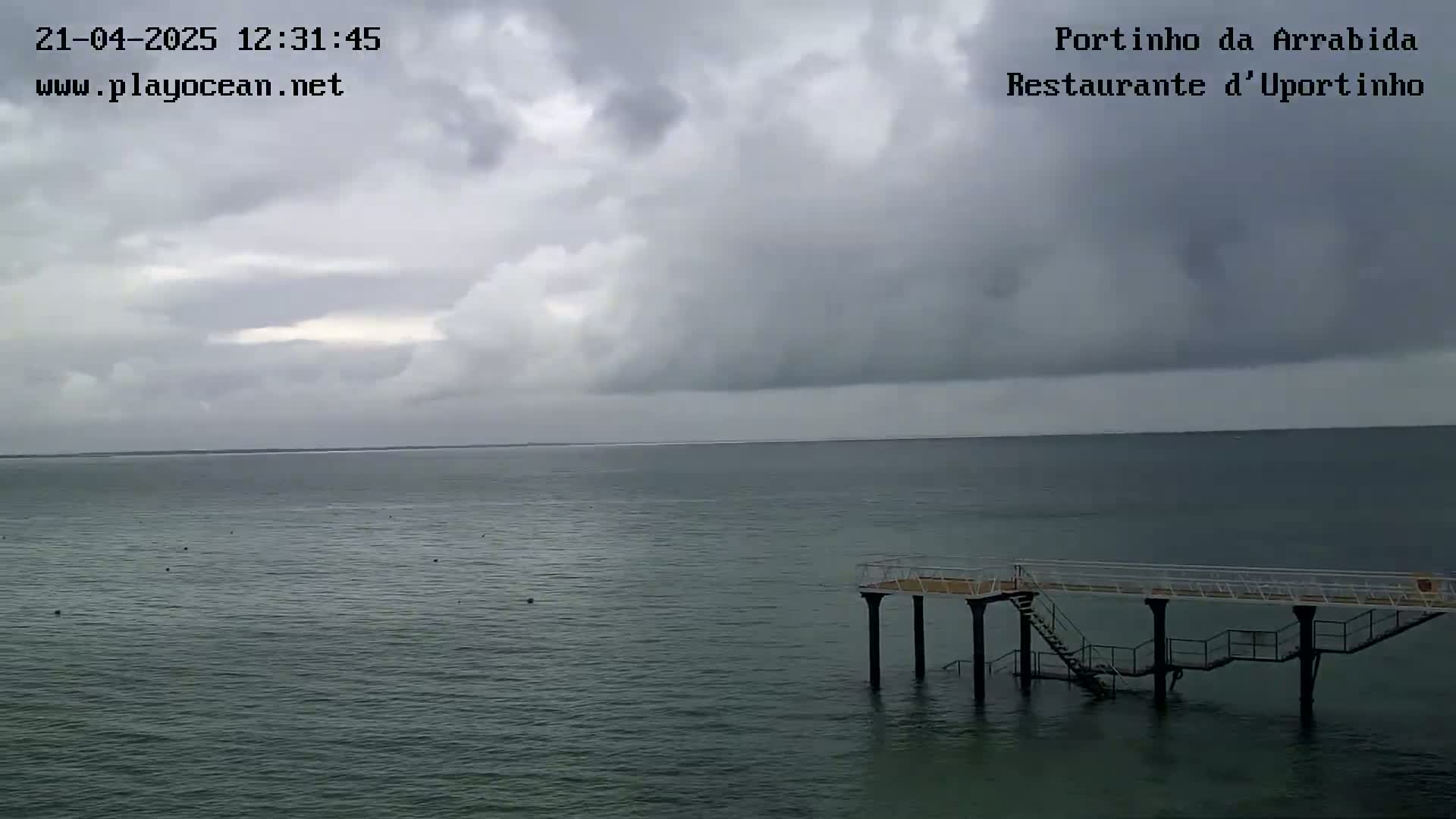 A dark, cloudy sky hangs over a calm, gray-green ocean, where a small pier with stairs extends into the water.