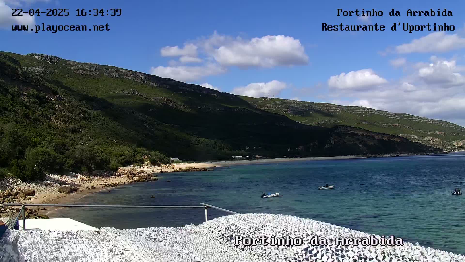 A partly cloudy blue sky overlooks a secluded cove with a sandy beach, green hills, and several small boats in the calm, clear water.