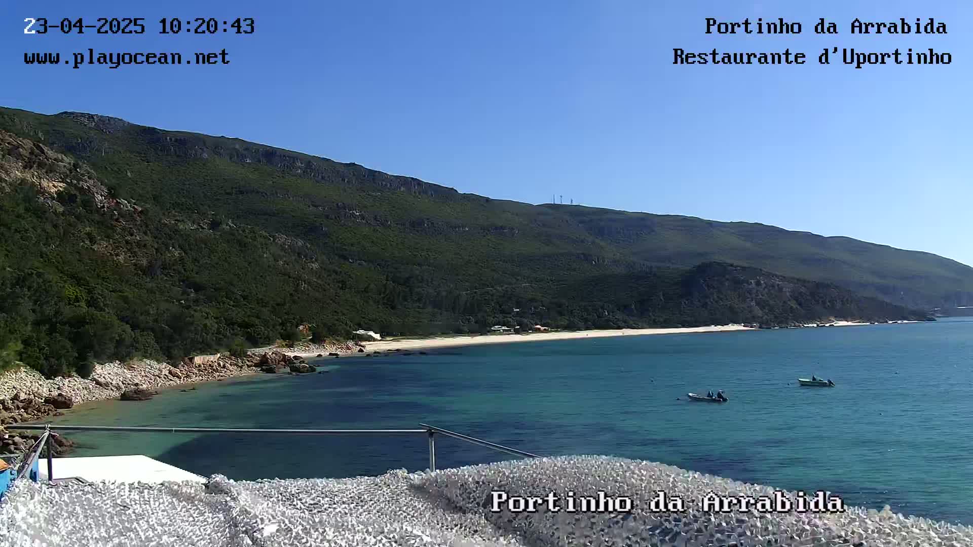 A tranquil beach cove with clear blue water is nestled beneath a lush green mountain on a sunny day.