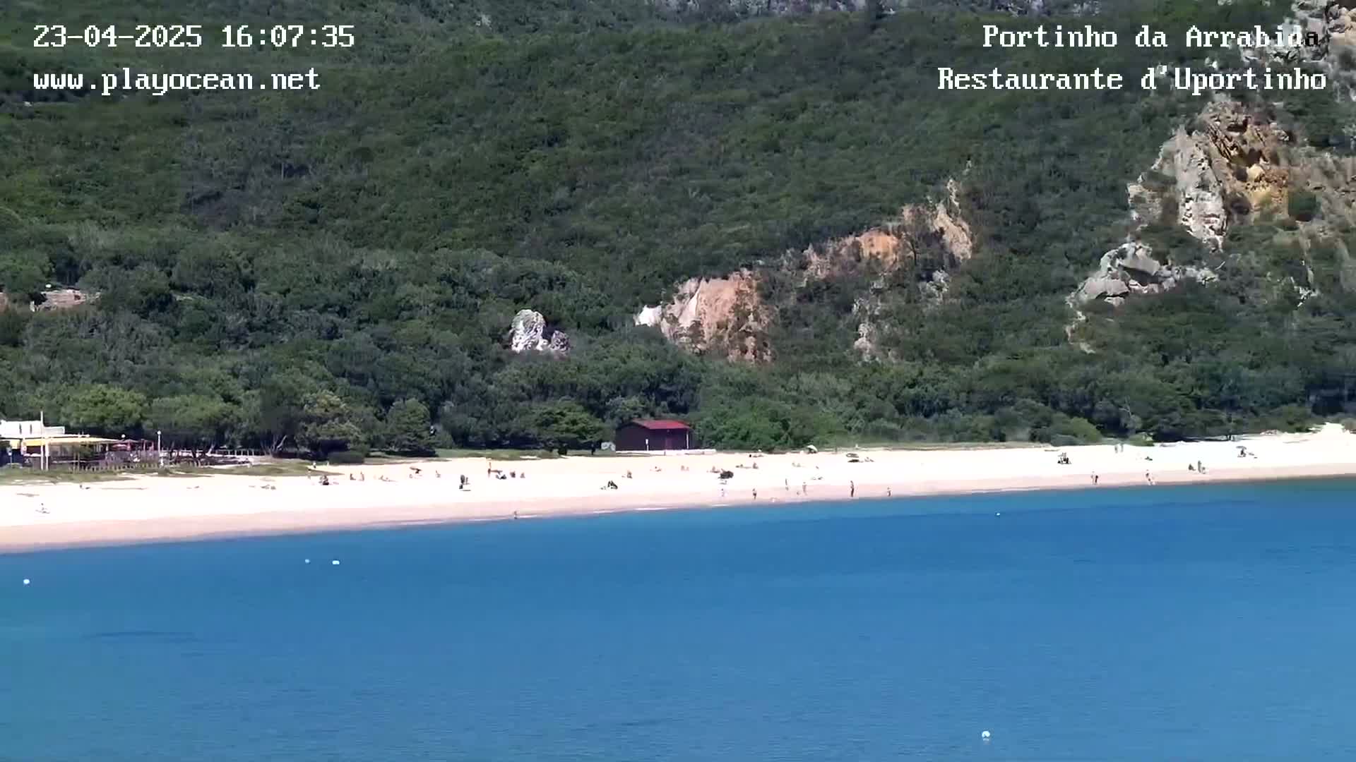 A sandy beach with a few people on it, backed by lush green vegetation and a small building, is situated next to calm, clear blue water.