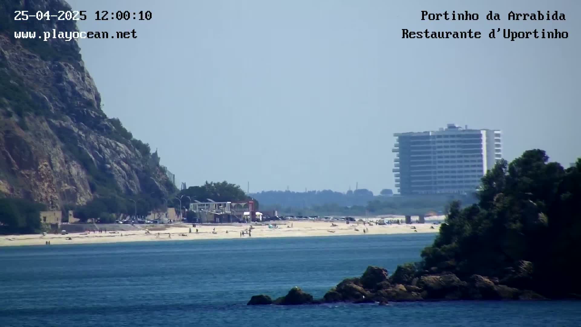 A sandy beach with scattered people is visible below a rocky cliff and a tall building, all under a hazy sky.