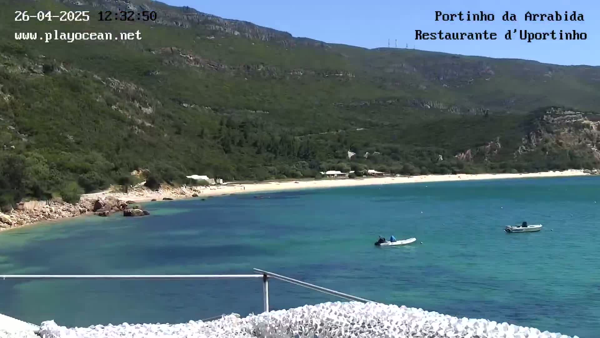 A sandy beach curves into a calm, clear turquoise bay under a sunny sky, backed by green hills and a few small boats.