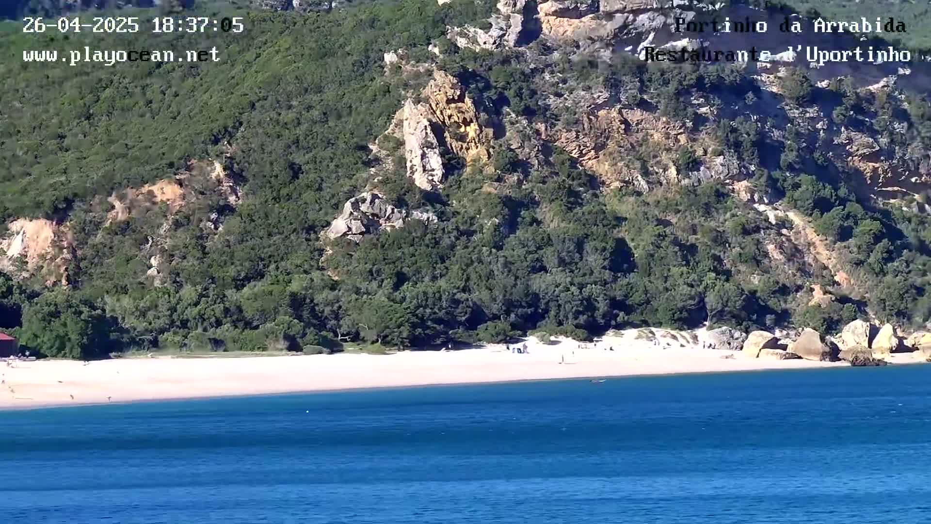 A sandy beach with a few people on it is nestled between green hills and a calm blue ocean.