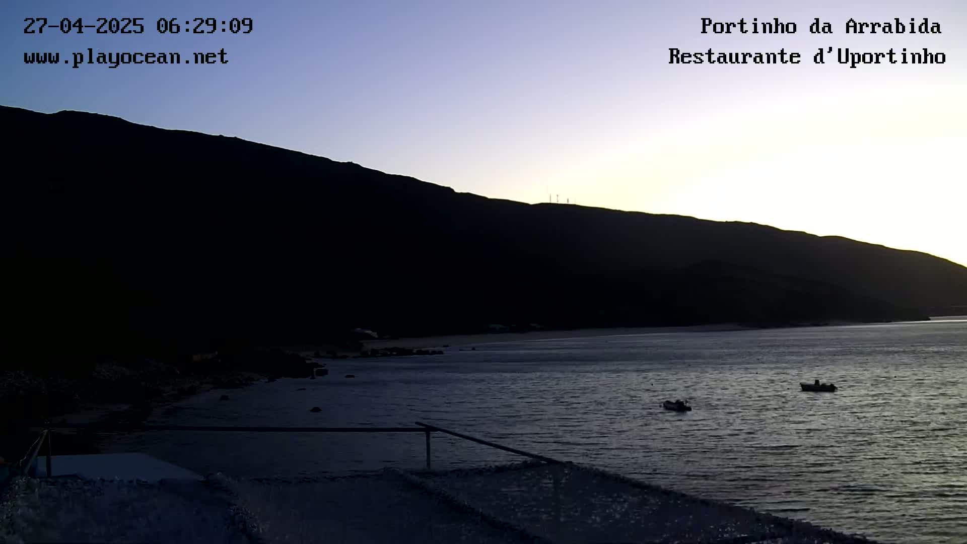 A dark, calm bay at dawn is nestled beneath a silhouetted mountain, with two small boats visible on the water.