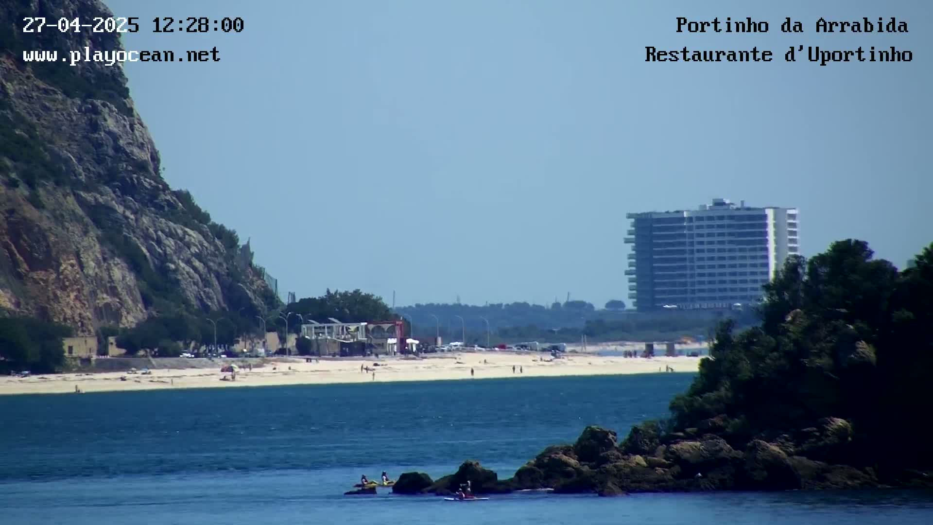 A sandy beach with a few people on it stretches between a rocky cliff and a tall building, with several people on jet skis in the calm blue water in the foreground.