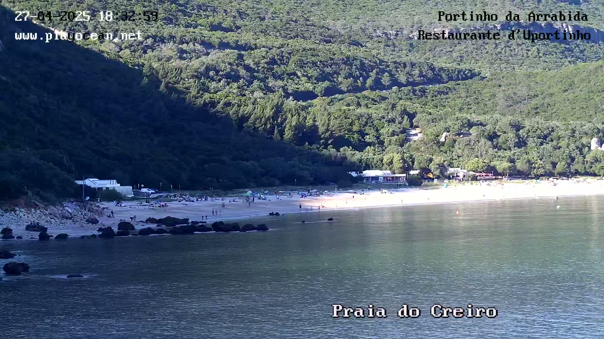 A sandy beach with people on it sits below a lush green hillside, next to calm, blue water.