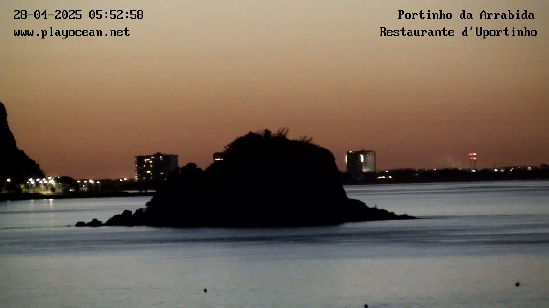 A dark, silhouetted island sits in calm water under a dusky orange sky at twilight, with city lights visible in the distance.