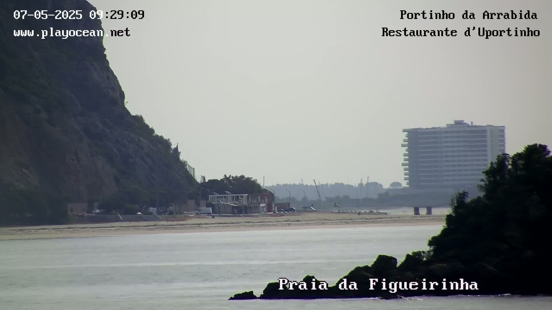A hazy view shows a beach with buildings and a cliff in the background, including a tall building in the distance.