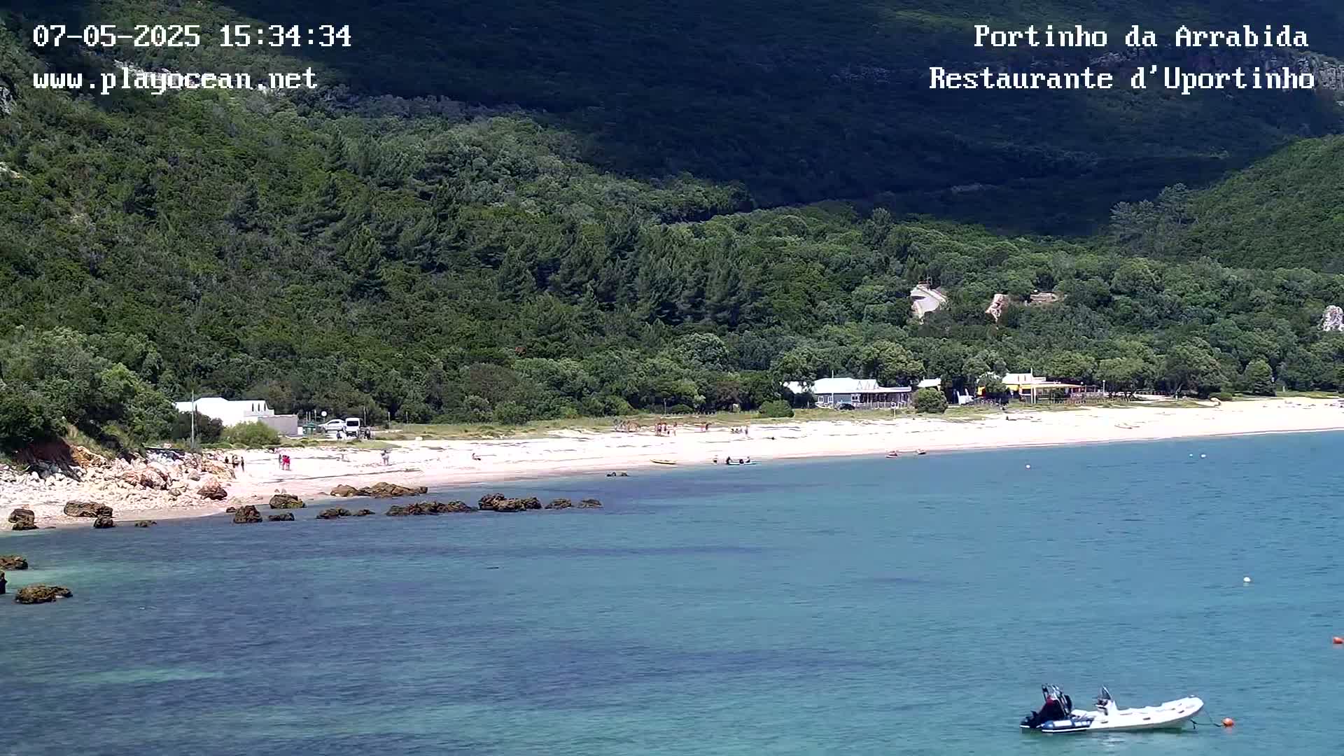A sandy beach with several people on it is nestled between a lush green hillside and calm, clear turquoise water, where a small inflatable boat is visible.