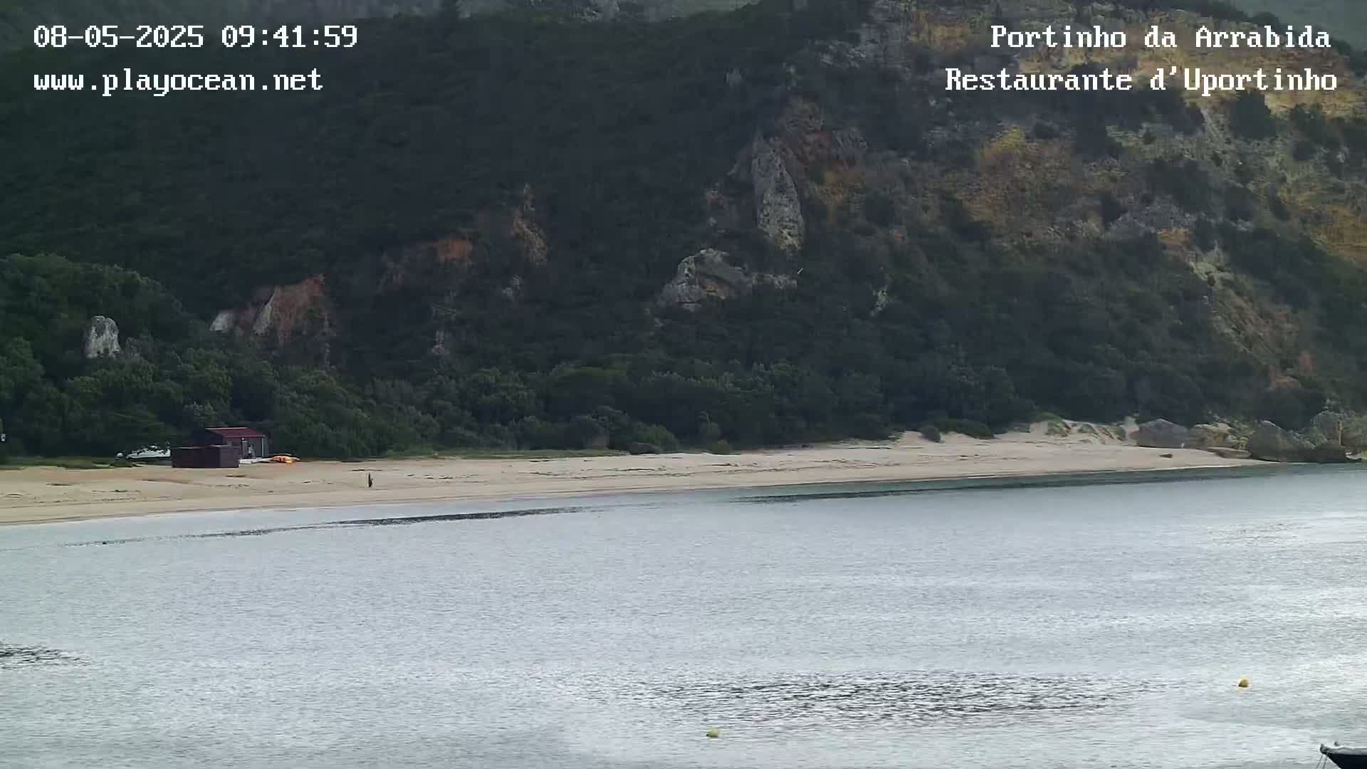A calm bay with a sandy beach backed by a rocky, tree-covered hillside.