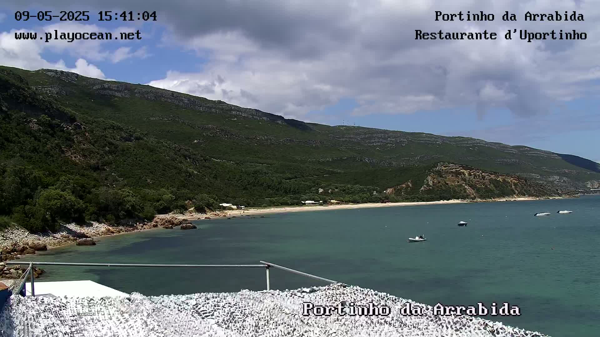 A partly cloudy day overlooks a tranquil cove with a sandy beach, nestled against a verdant mountainside, where several small boats are visible in the calm, teal water.