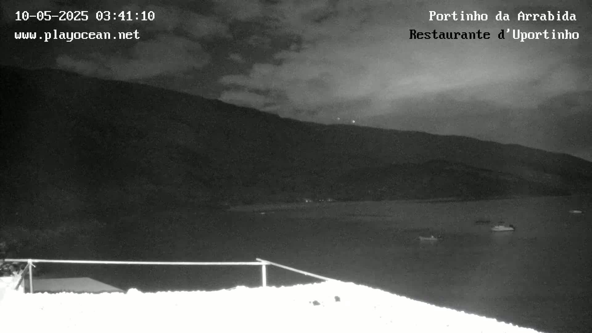 A nighttime, low-light view shows a dark body of water with a few boats, in front of a dark mountain under a cloudy sky.