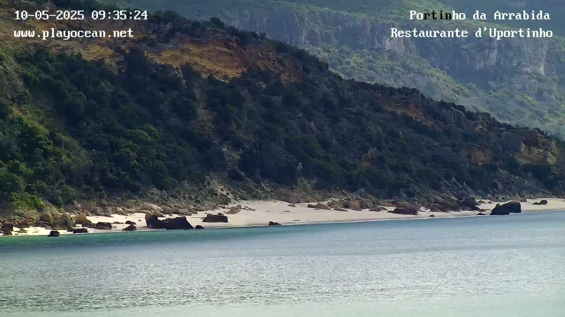 A tranquil beach with light-colored sand and dark rocks sits at the base of a lush, green cliff on a calm, sunny day.
