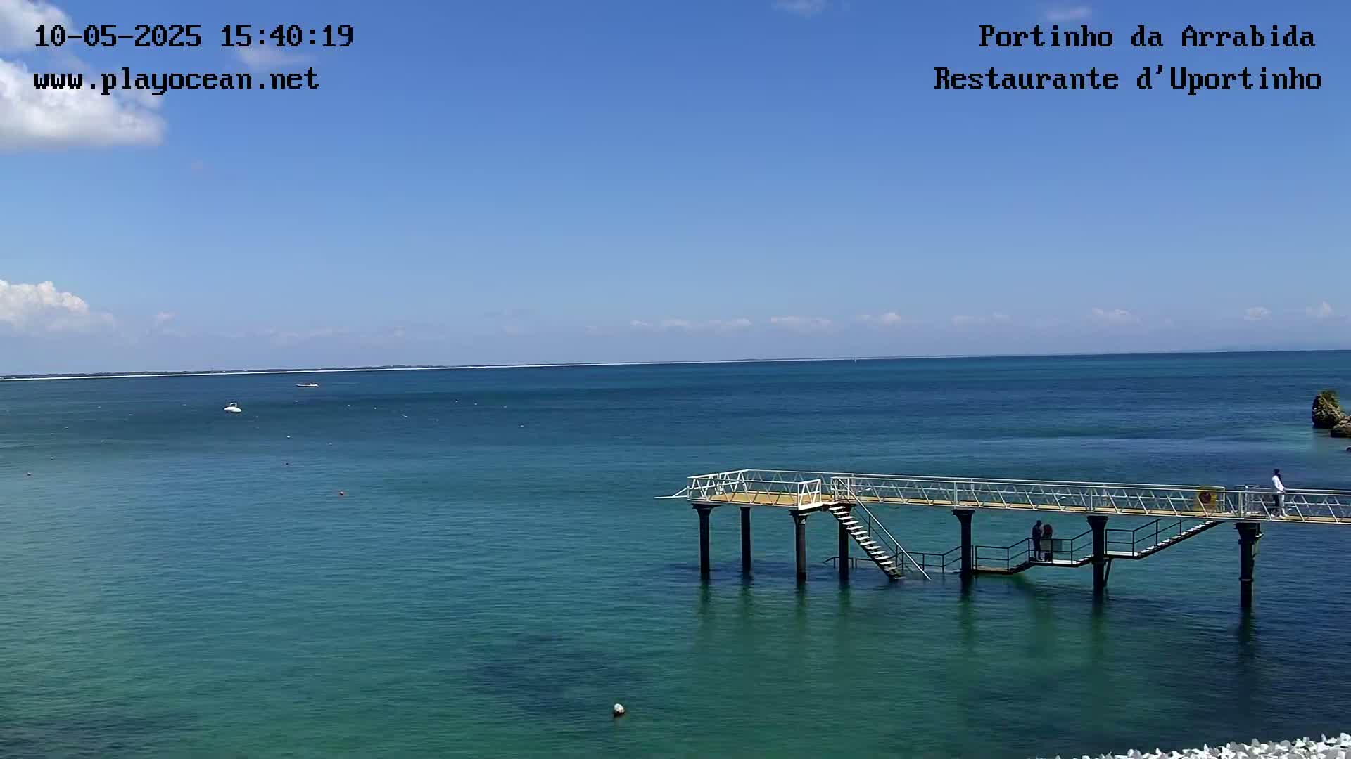 A metal pier extends over a calm, teal ocean under a mostly clear blue sky with a few small clouds.