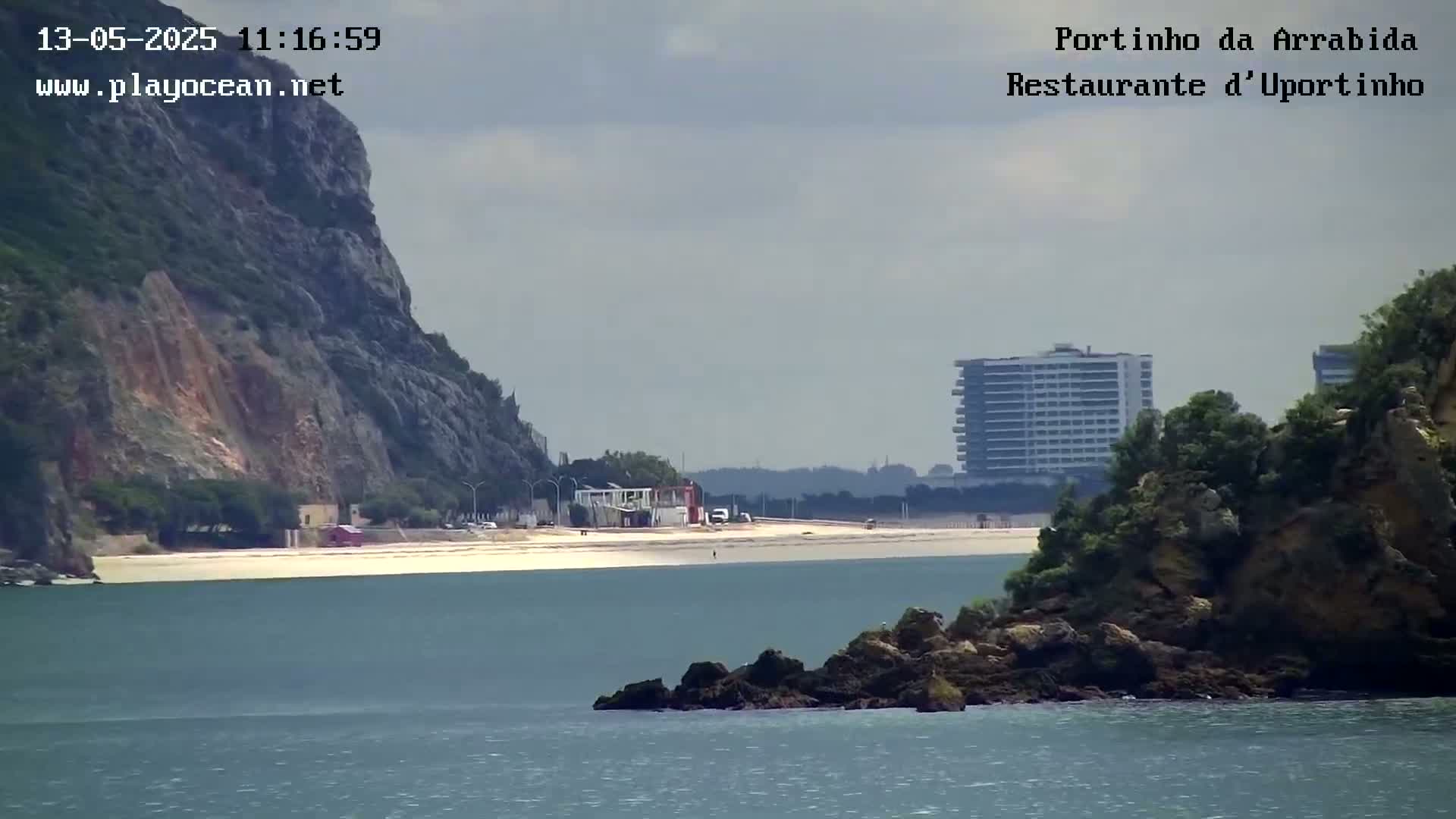 A sandy beach sits between a rocky cliff and a distant high-rise building under a partly cloudy sky.