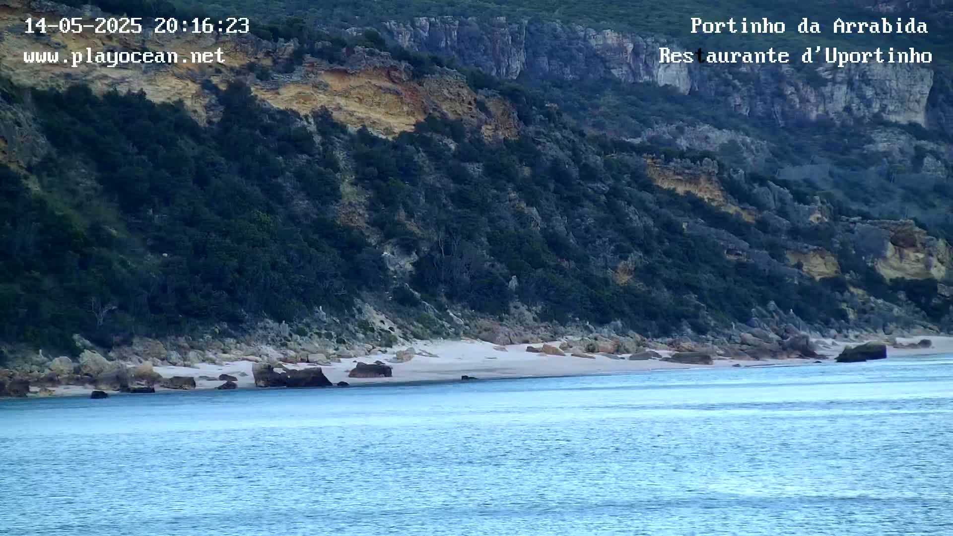 A sandy beach sits below a dark green, rocky cliffside overlooking calm blue water.