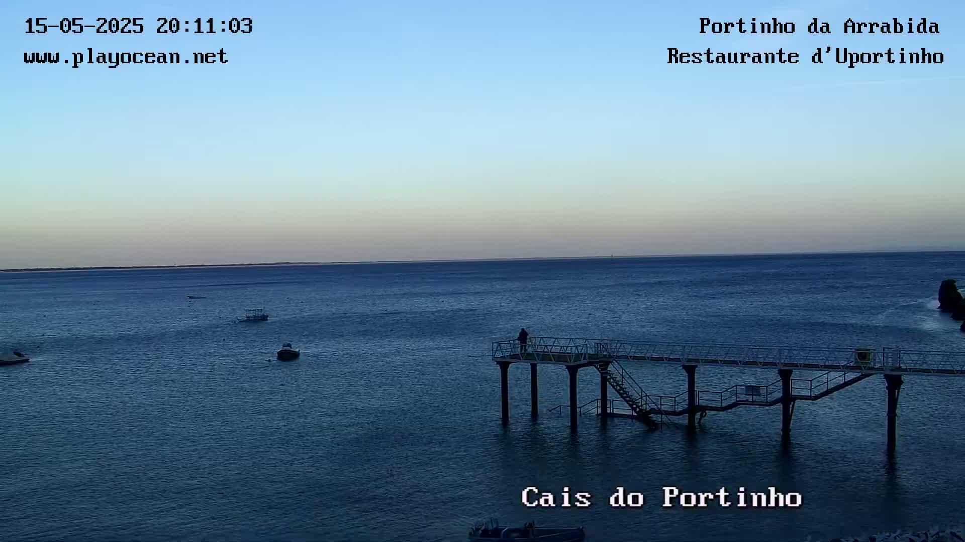 A calm, twilight ocean scene features a pier extending into the water with a lone figure standing on it, a few small boats are visible in the distance.