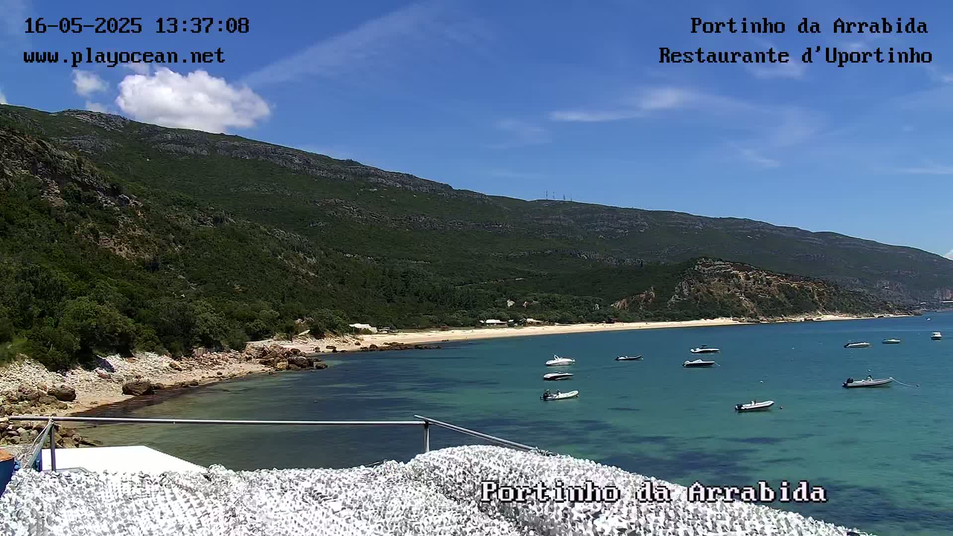 A secluded sandy beach with several small boats in clear, calm water is nestled beneath a verdant, mountainous landscape under a mostly sunny sky.