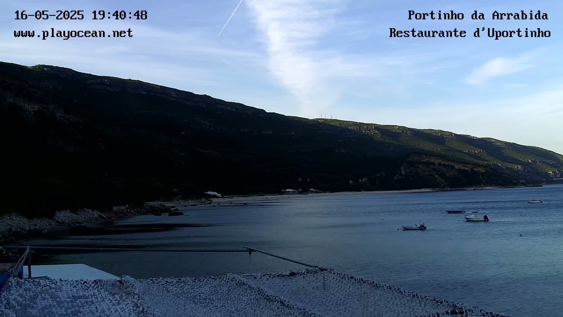 A calm bay with a few small boats is nestled beneath a dark, mountainous coastline under a mostly clear, twilight sky.