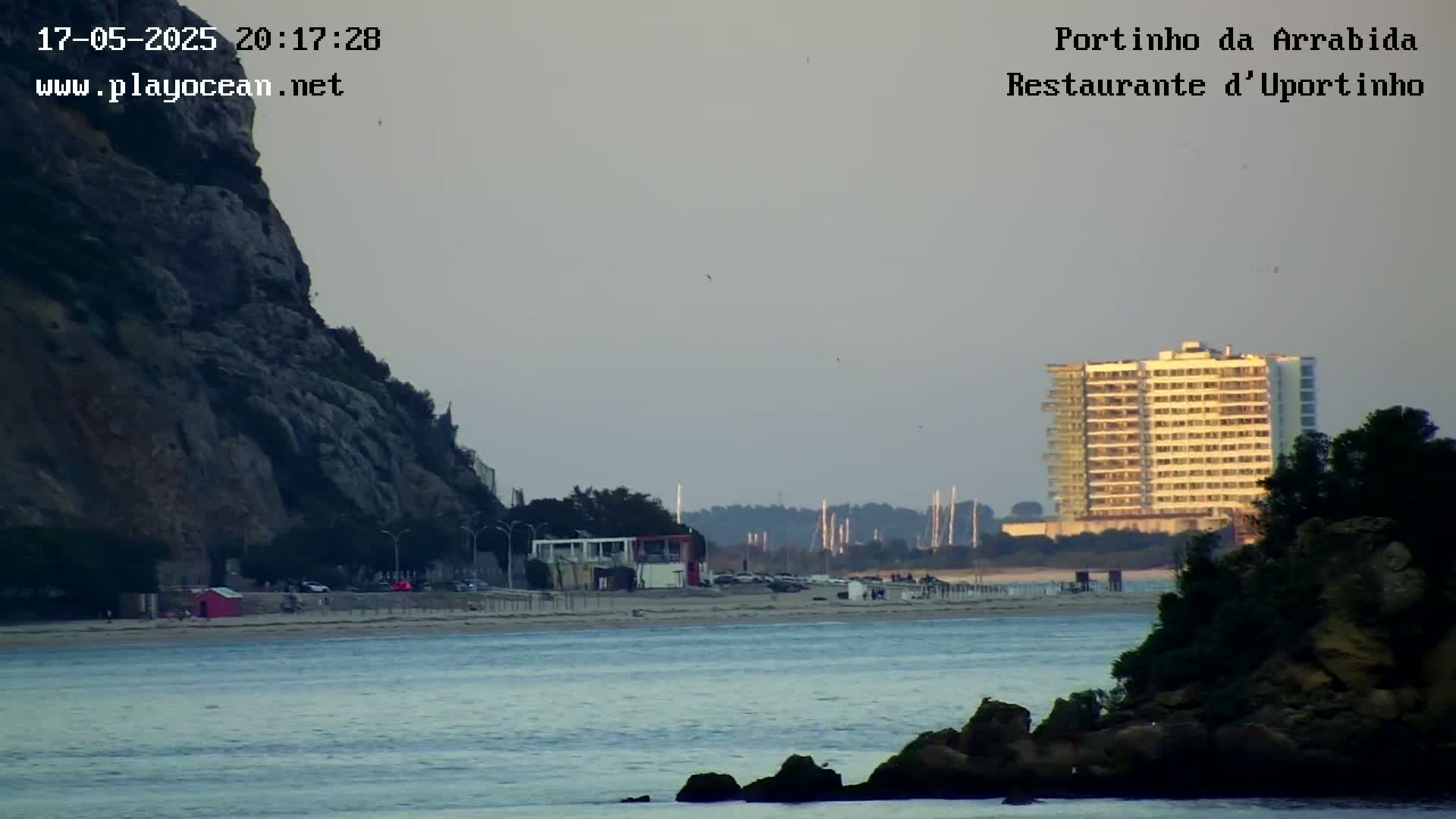 A beach scene at sunset shows a rocky cliff to the left, a sandy beach with buildings, a multi-story hotel in the distance, and rocky outcroppings in the foreground; the water is calm.