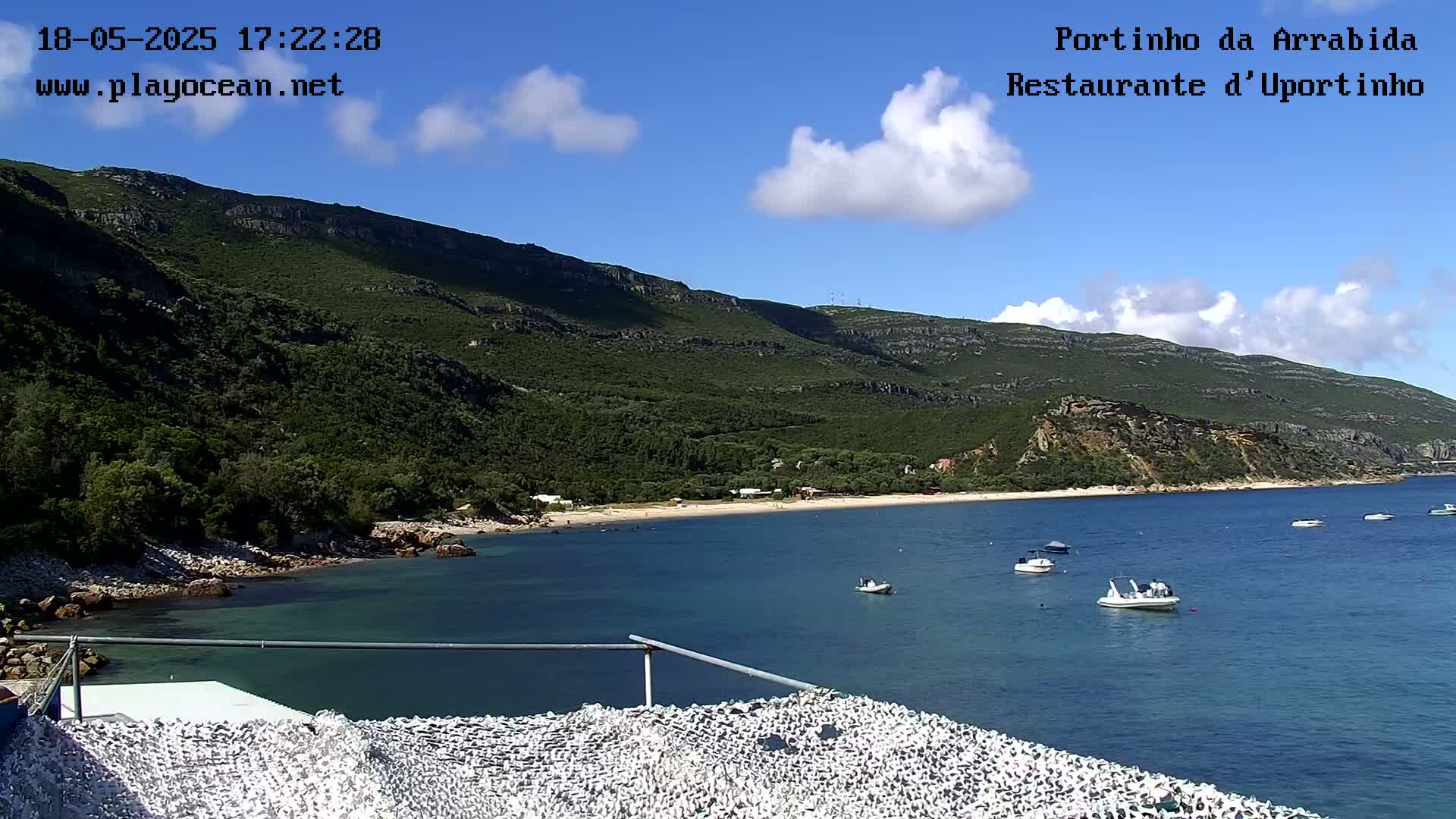 A calm bay with several small boats is nestled beneath a verdant mountain range under a mostly sunny sky with some scattered clouds.