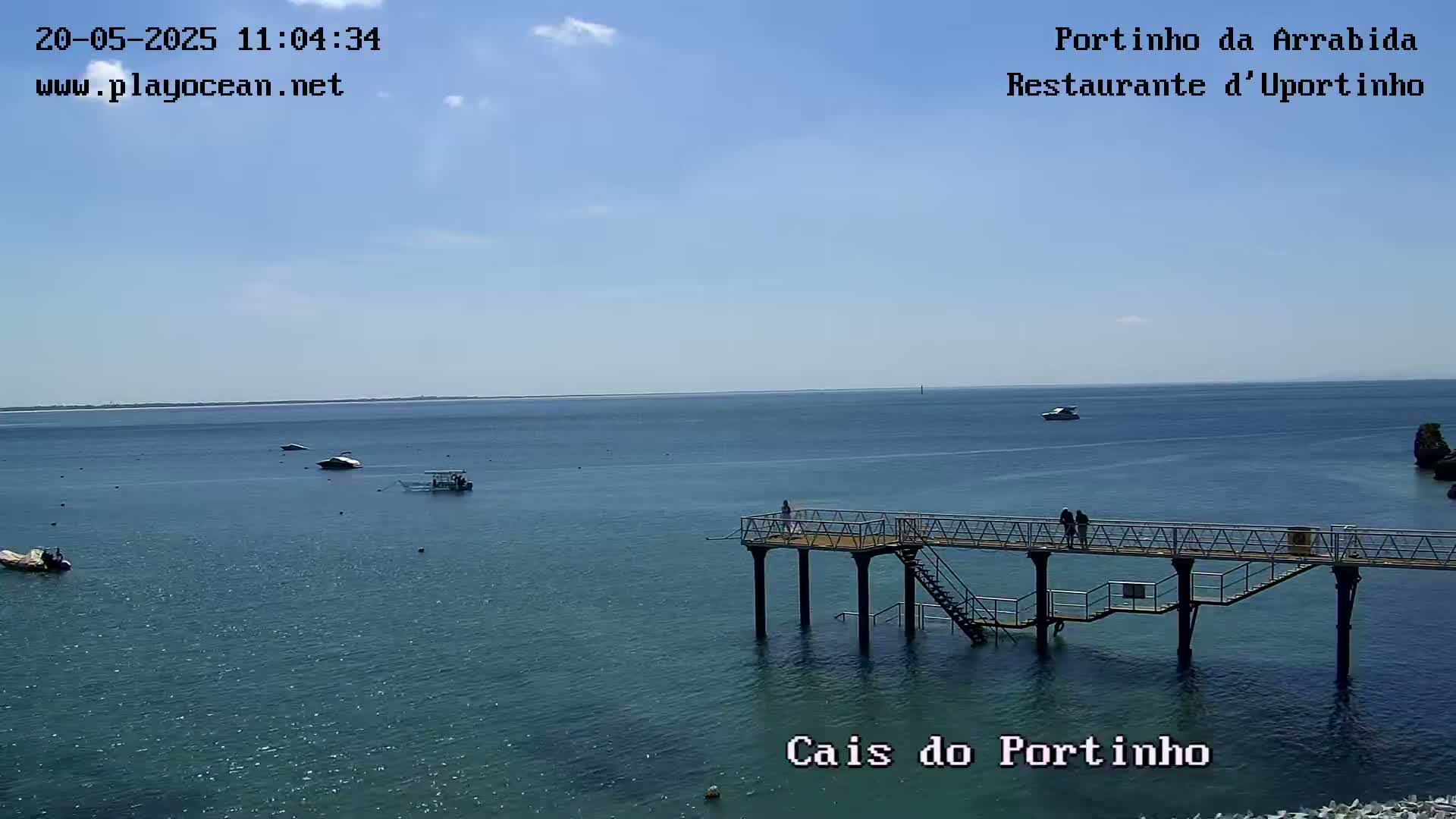 A pier extends over calm, blue water with several small boats visible in the distance under a clear, sunny sky.