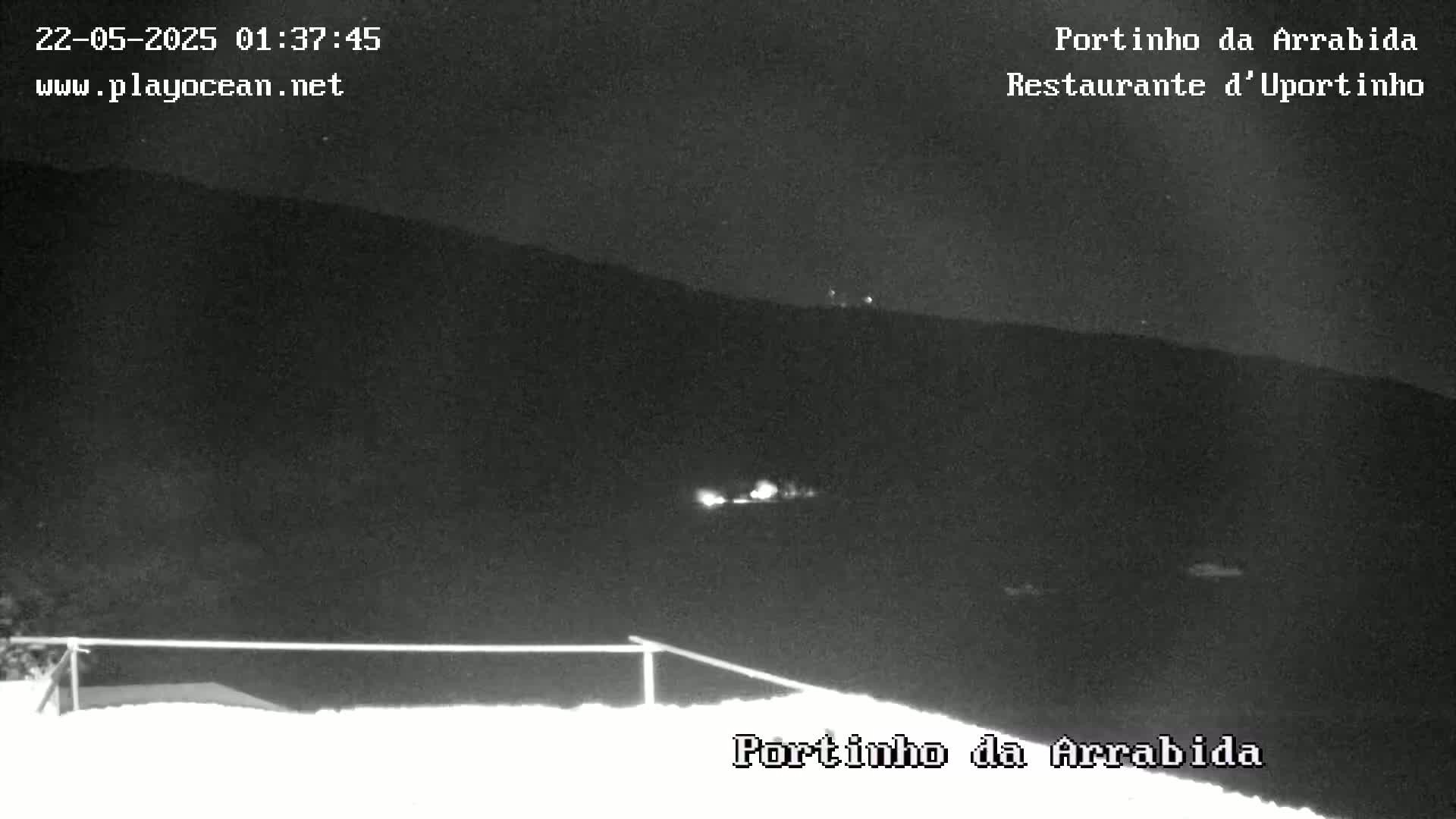 A nighttime, low-light image shows a dark landscape with a few distant lights visible, possibly indicating a small town or village, seen from a vantage point with a low railing in the foreground.