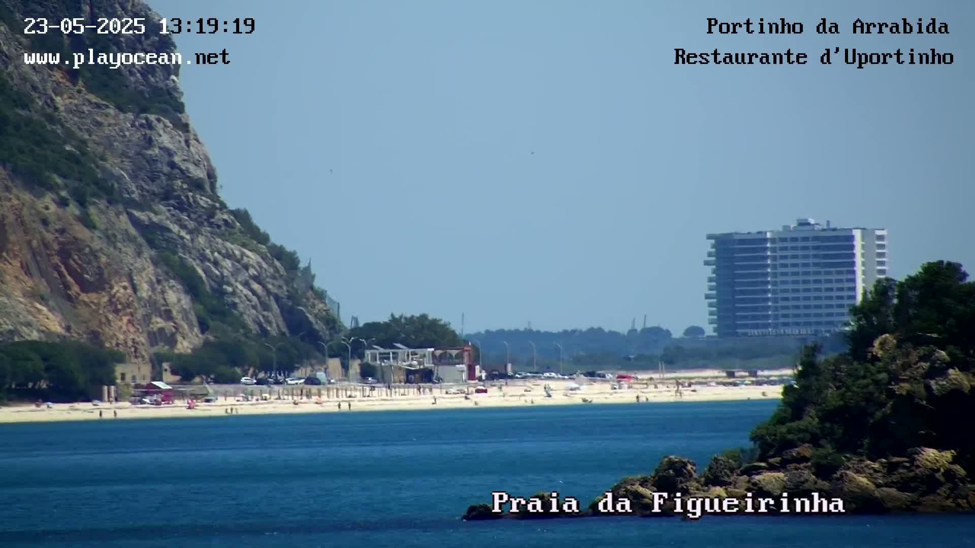 A sandy beach with people on it is visible from across a calm body of water, with a rocky cliff face on the left and a high-rise building in the distance under a clear sky.