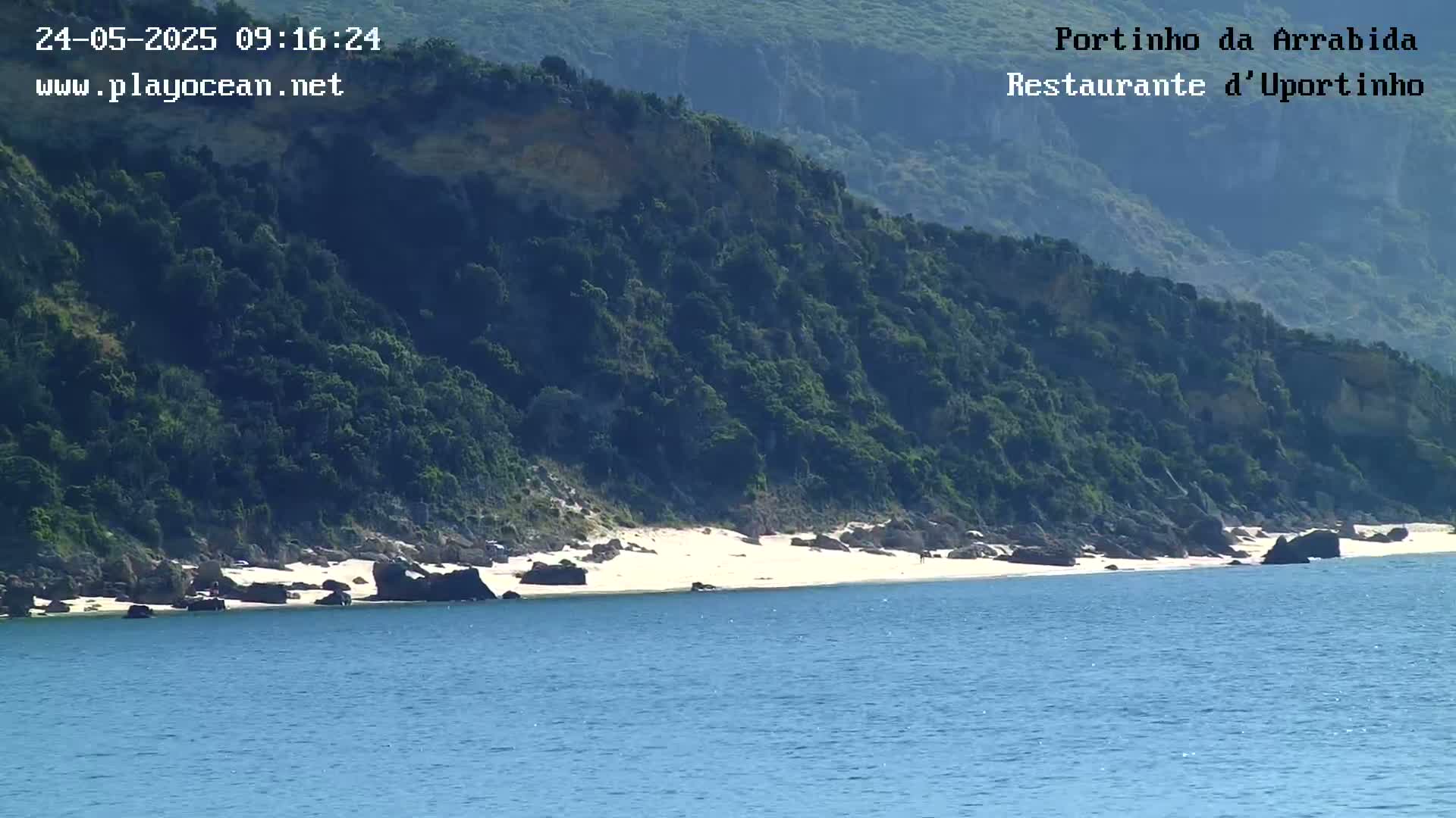 A small, sandy beach lies nestled between dark green, rocky hills and a calm, blue ocean on a sunny day.