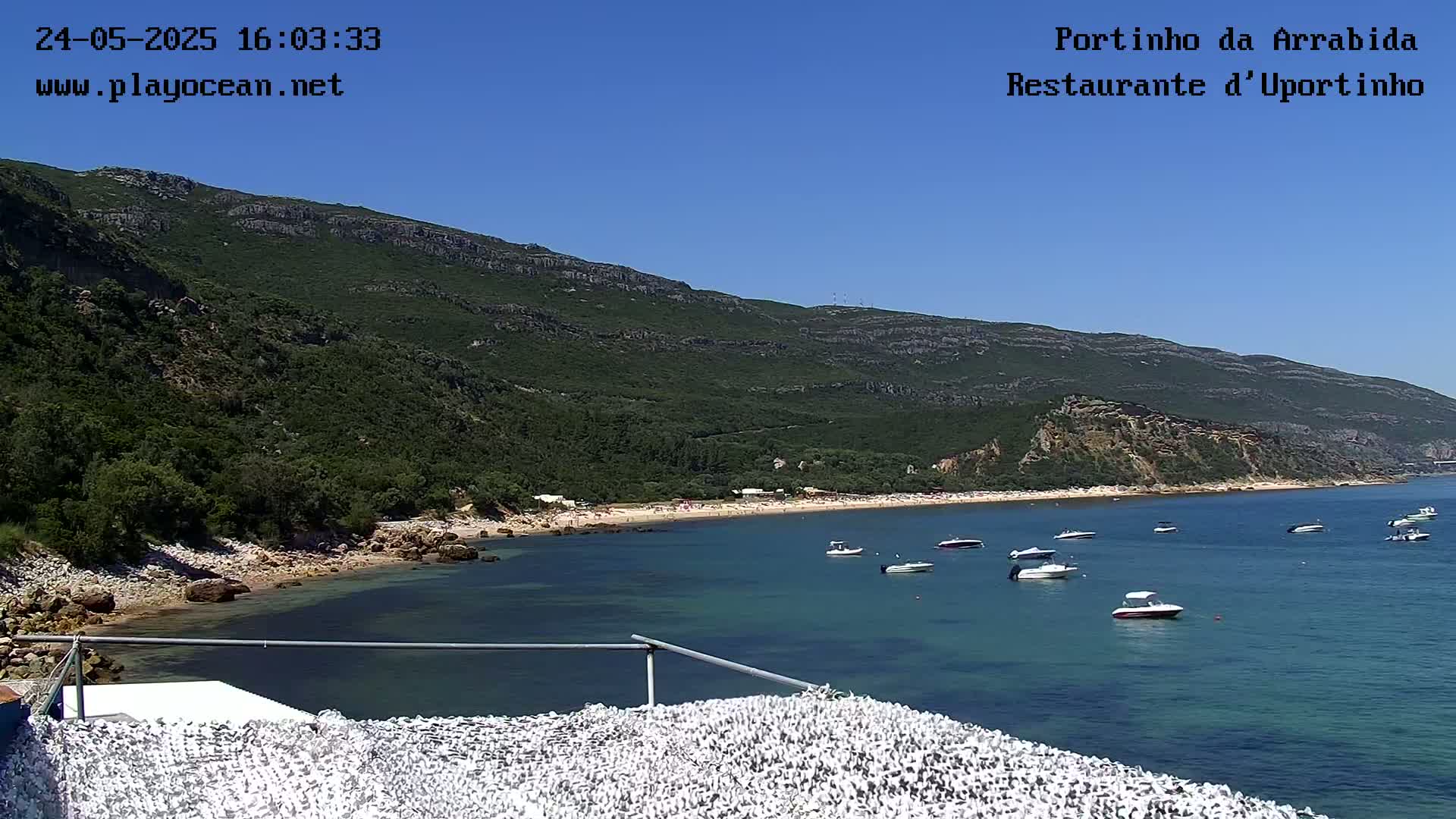 A sandy beach with several small boats in calm, clear blue water is nestled below a large, green mountain on a sunny day.