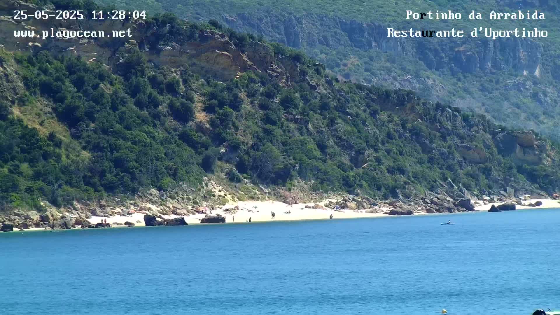 A sandy beach with a few people on it is nestled below a lush, green cliffside overlooking calm, blue water.