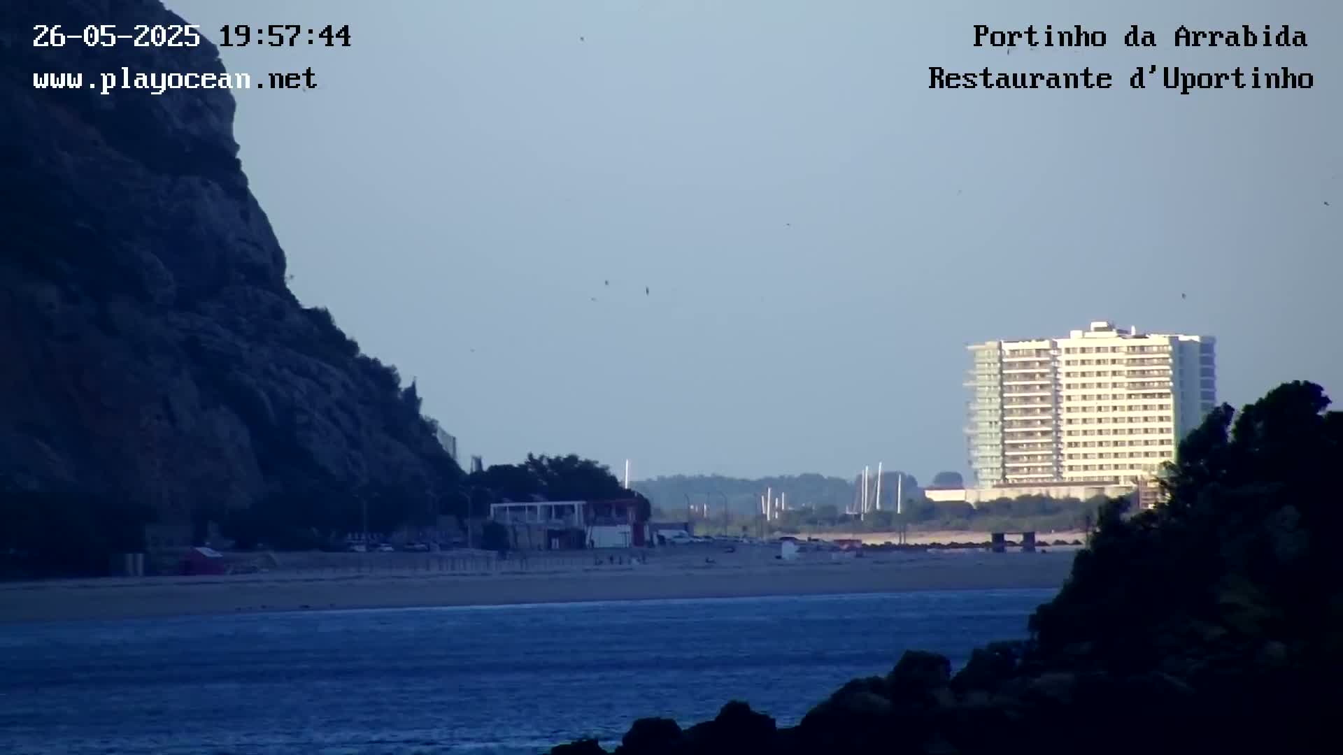 A sandy beach with calm water stretches between a rocky cliff and a tall building, under a clear, dusky sky.