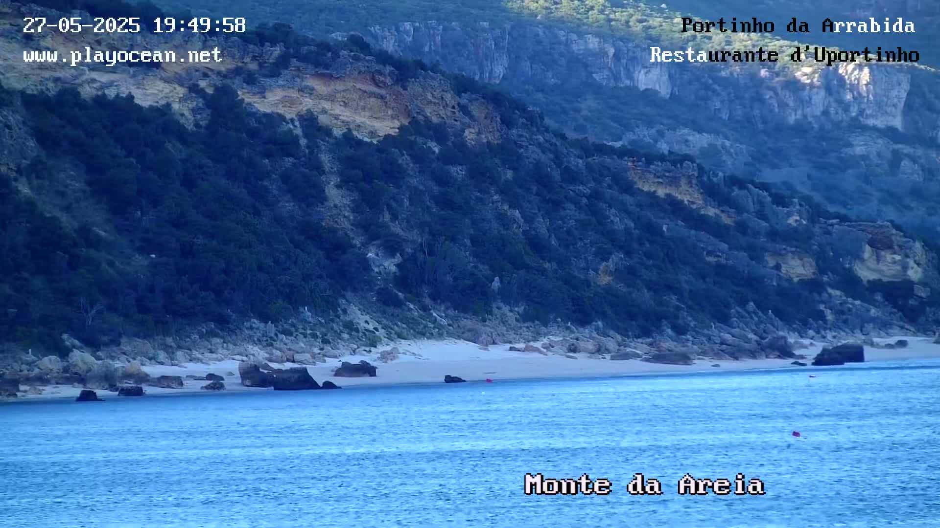 A tranquil beach with light-colored sand is nestled between a dark-green, rocky cliff and a calm, blue ocean.