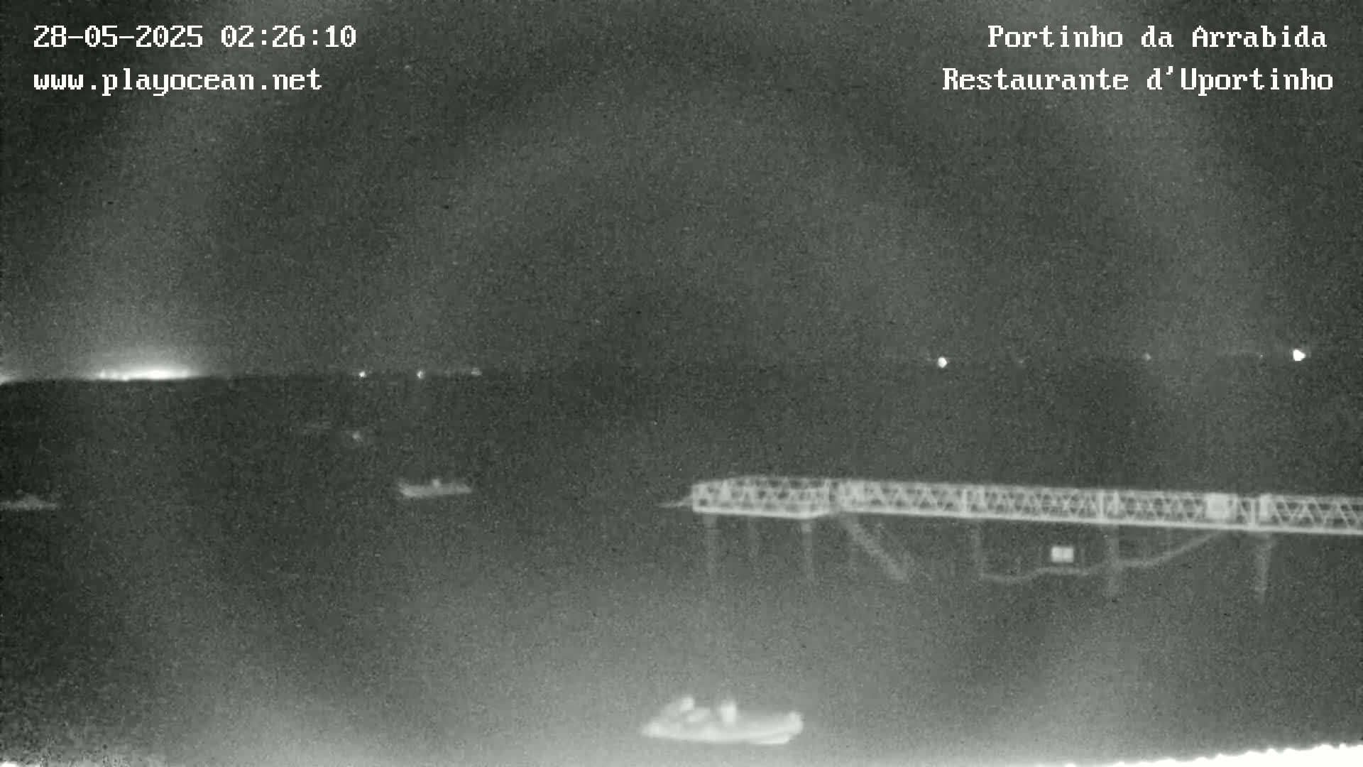 A nighttime, low-light image shows a pier extending into dark water, with a few small boats visible, and a faintly lit horizon in the distance.