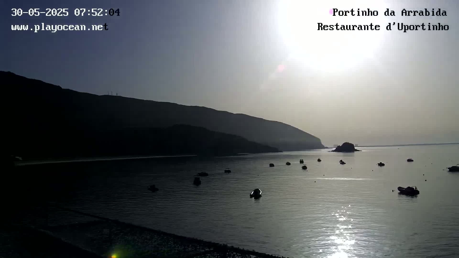 A calm body of water with several small boats is nestled in a bay, partially shaded by a dark mountain under a bright, hazy sun.