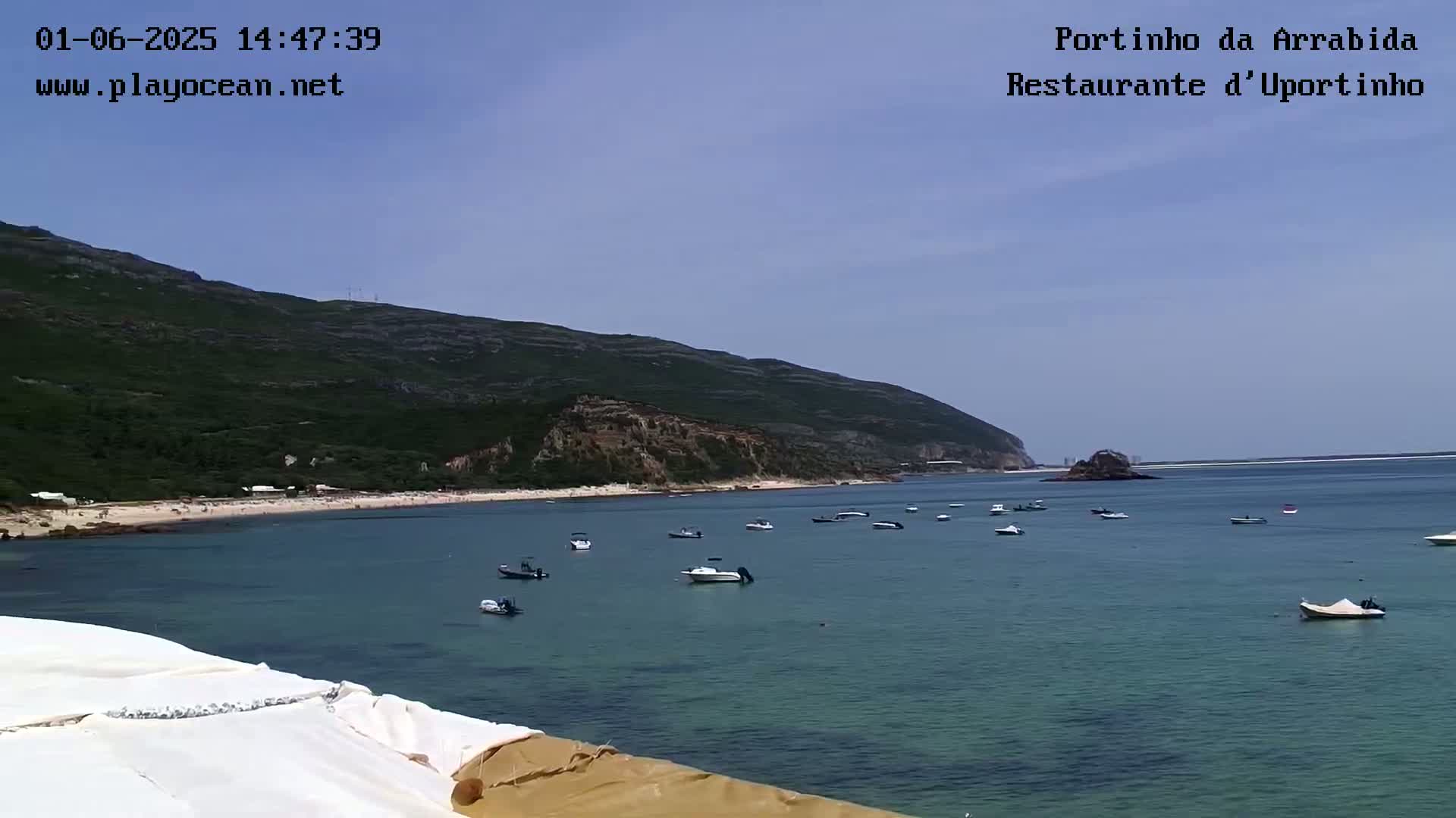 A calm, clear day reveals a bay with numerous small boats, a sandy beach, and a green mountain in the background.