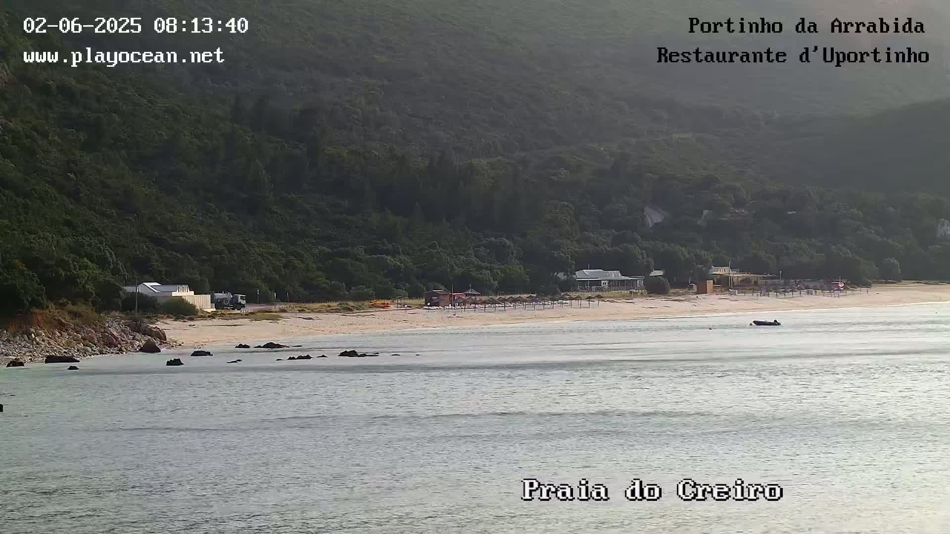A sandy beach with a few buildings and beach umbrellas is nestled between a calm body of water and a lush, green hillside.