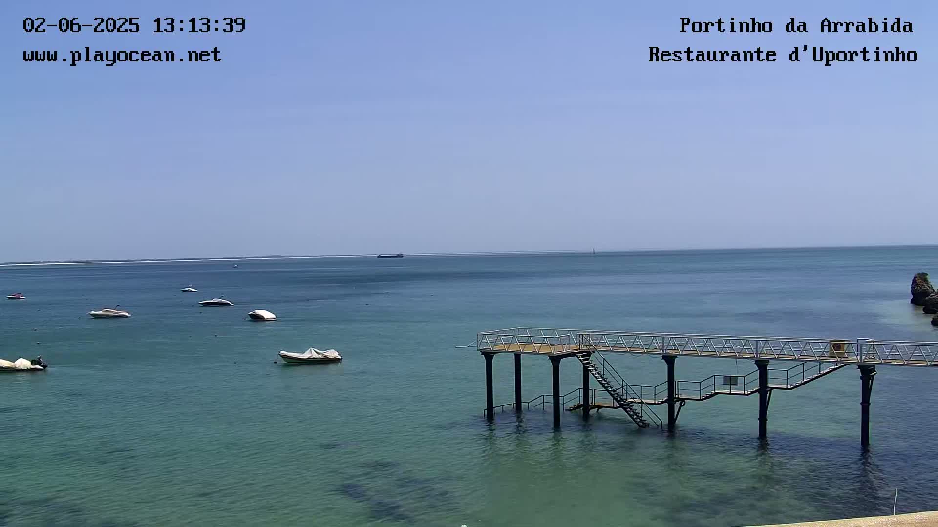 A calm, clear day shows a body of water with several small boats and a long pier extending into the sea.