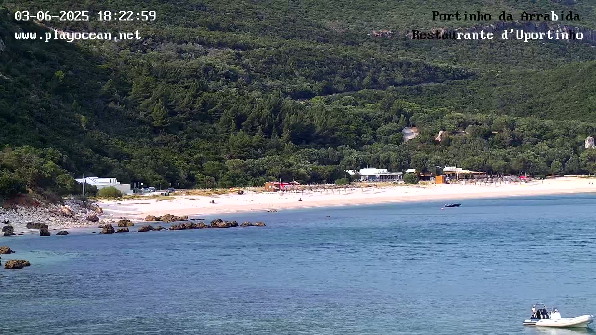 A sandy beach with calm, blue water, backed by green hills and a few small buildings, is seen on a sunny day.