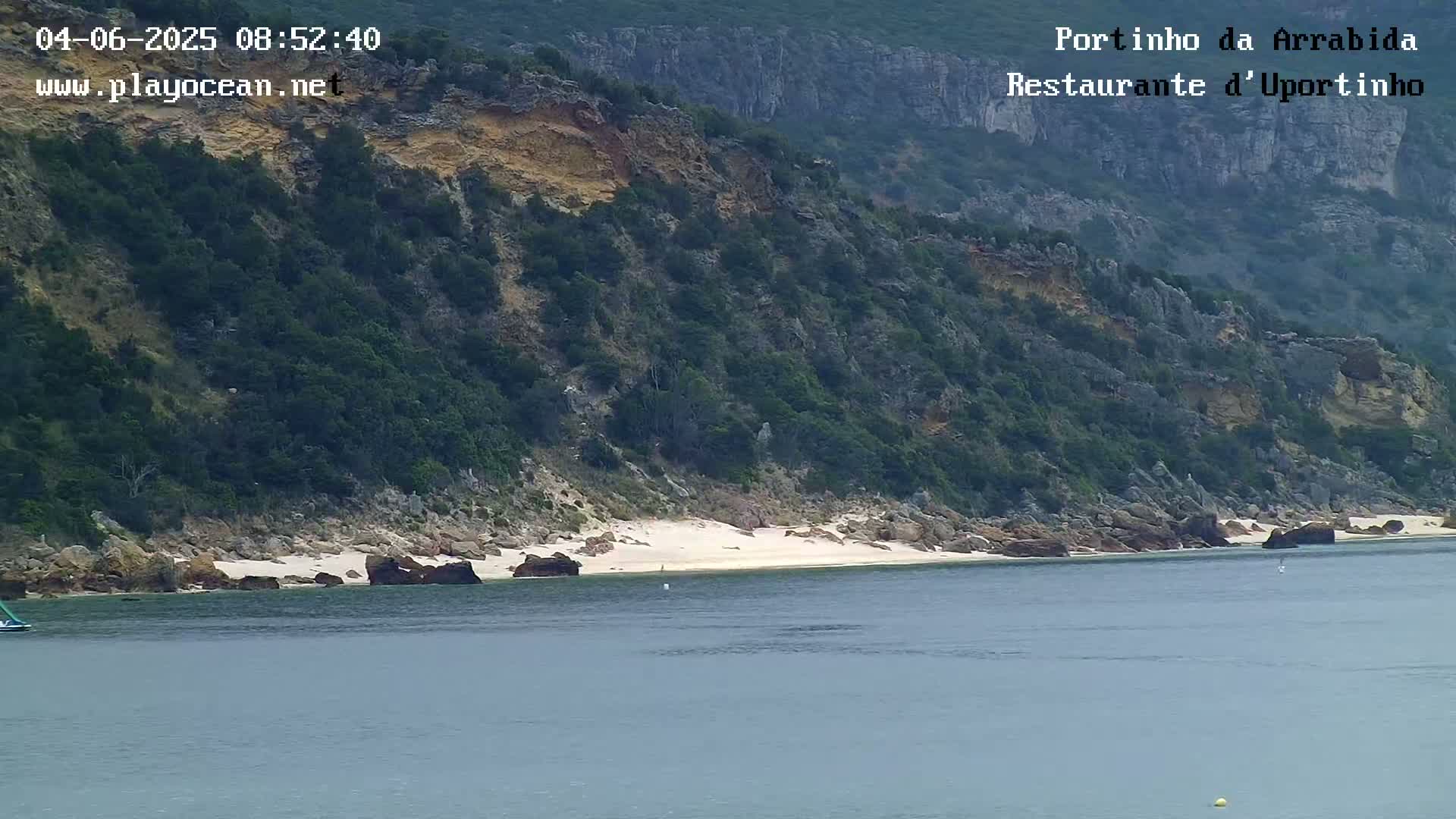 A secluded, sandy cove nestled between rocky, green-covered hills meets a calm body of water under an overcast sky.