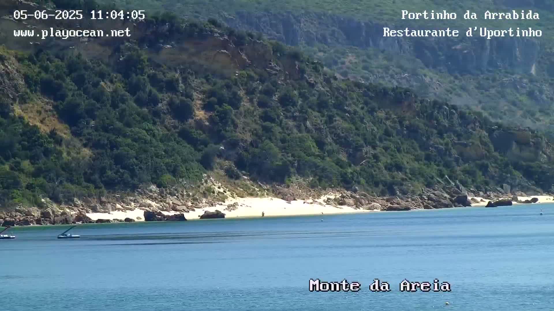 A tranquil beach with calm water is nestled between a rocky, lush green hillside and a calm blue ocean under a clear sky.