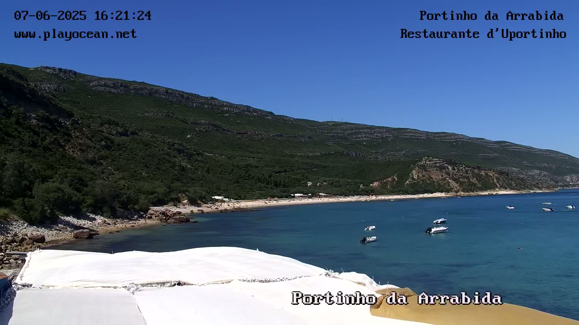 A sandy beach curves around a calm, blue bay under a clear blue sky, backed by a lush green mountainside.