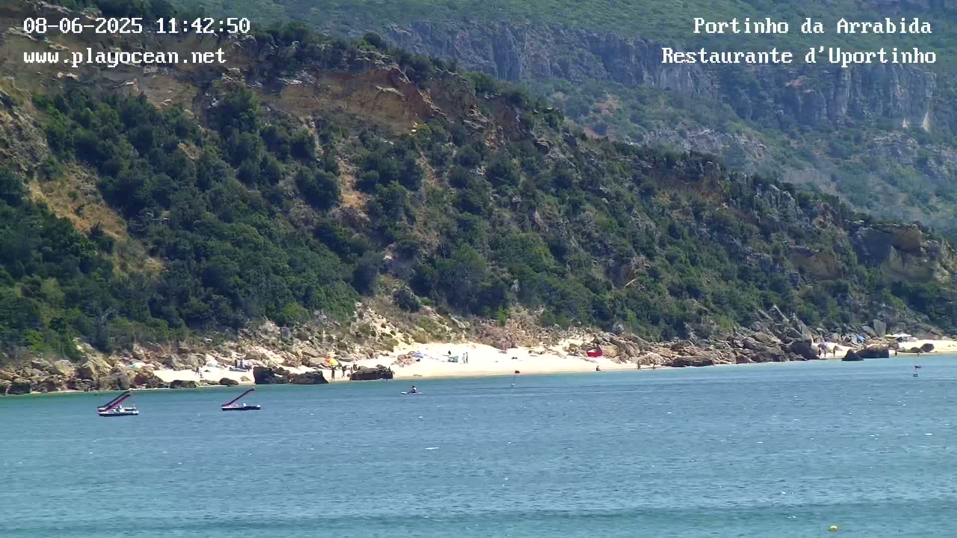 A sandy beach with several people on it sits at the base of a rocky, green hill overlooking calm blue water with two small boats.