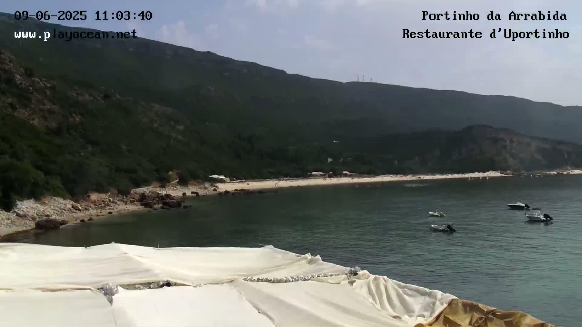 A sandy beach nestled in a cove below a verdant hillside, with several small boats in the calm water on a partly sunny day.