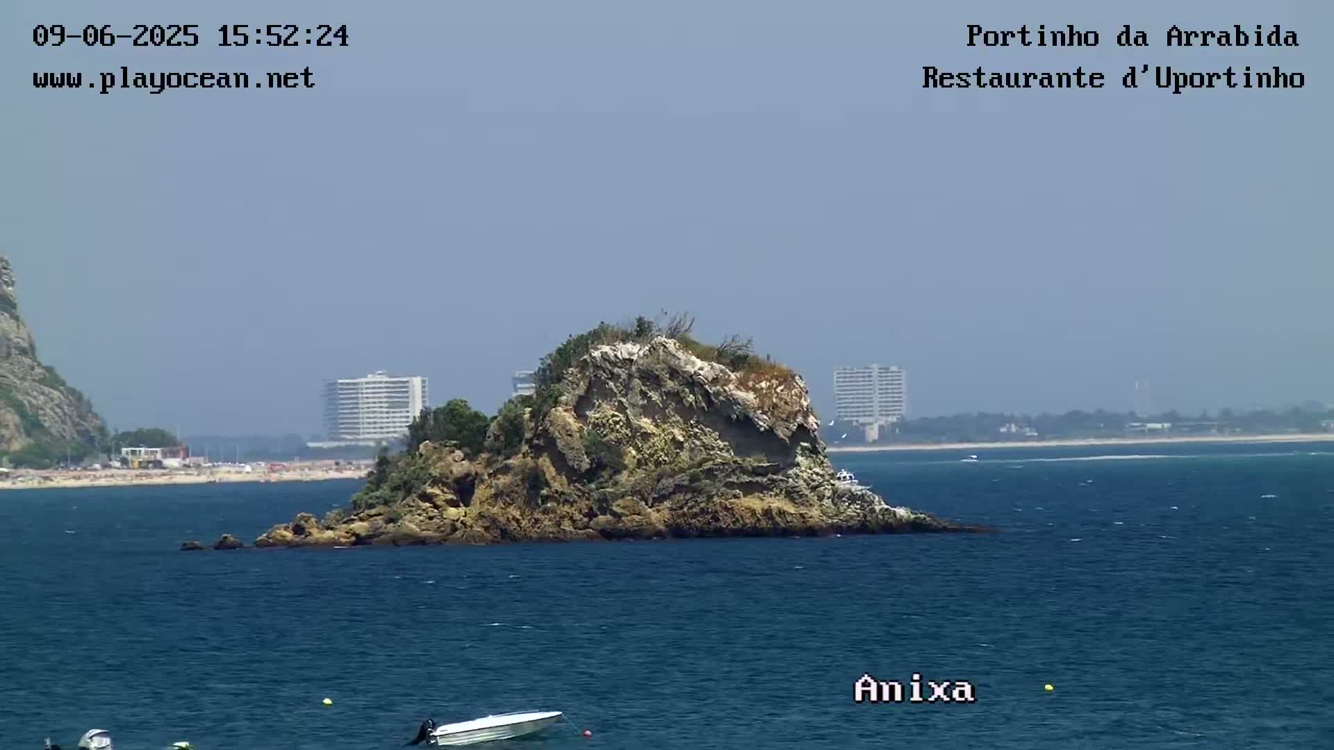 A rocky island sits in a calm blue ocean under a clear sky, with a sandy beach and buildings visible in the distance.