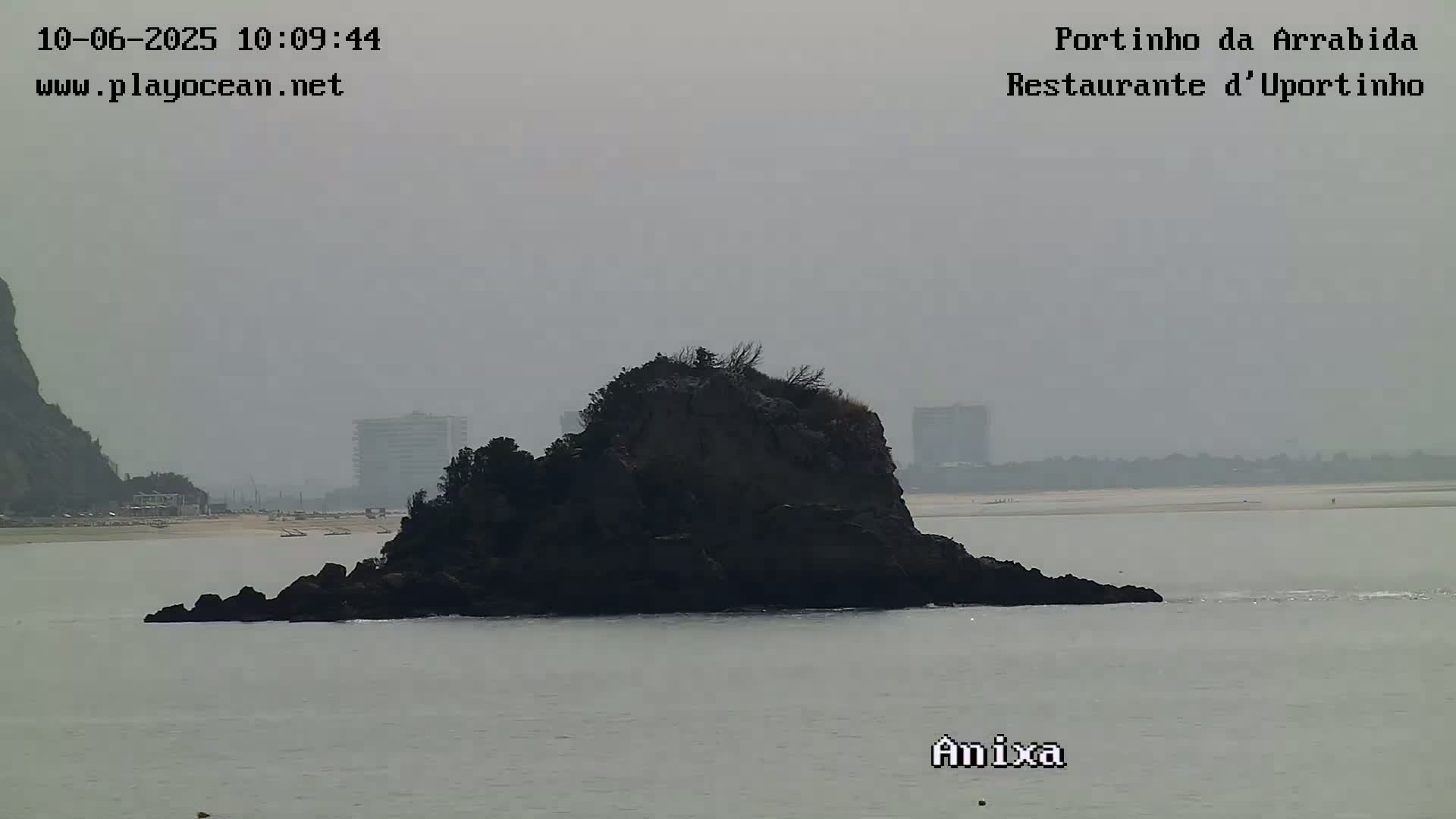 A hazy view shows a dark rock formation in calm water, with a sandy beach and distant buildings visible in the background.