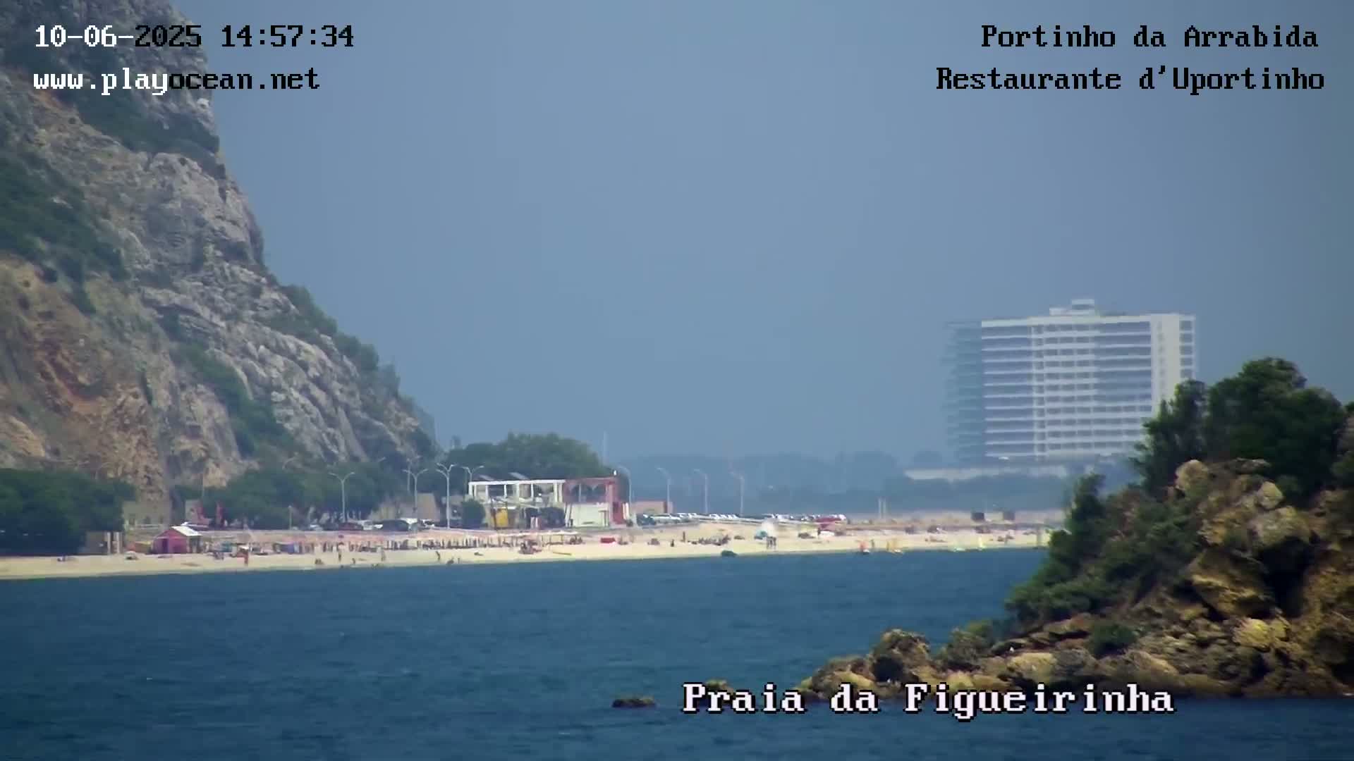 A hazy view shows a beach with people on it, backed by a cliff, buildings, and vegetation, all under a mostly clear sky.