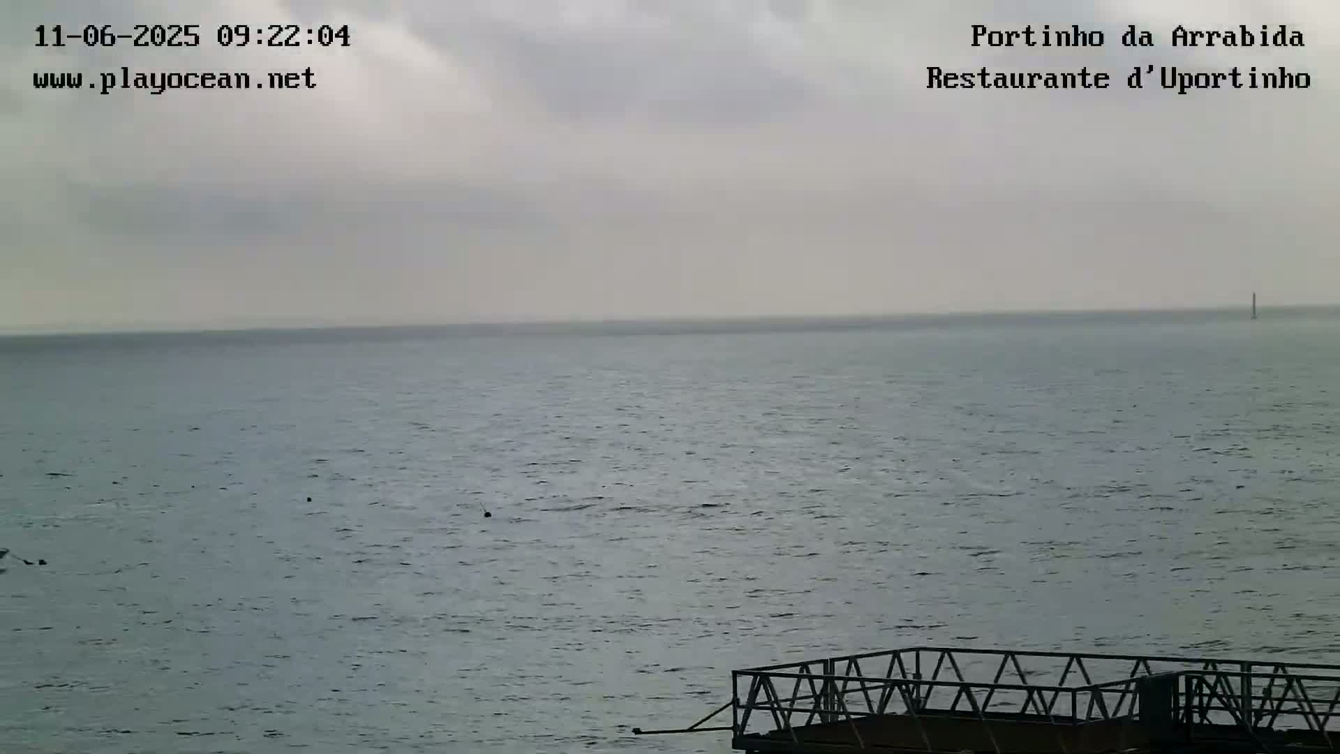 A mostly cloudy day reveals a calm, gray-blue ocean with a small metal pier in the foreground.